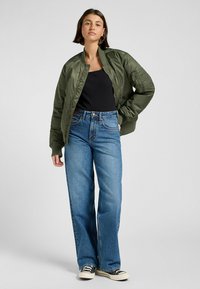 Olivgrüne Bomberjacke mit gerippten Bündchen, kombiniert mit hellblauen weit geschnittenen Jeans und einem schwarzen Top. Trägt legere Sneaker mit weißen Sohlen.