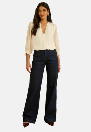 Sieviete ar gariem tumšiem matiem, valkā krēmkrāsas blūzi ar pūderētām piedurknēm, tumšas plataji džinsas un burgundijas krāsas norādītas kurpes.