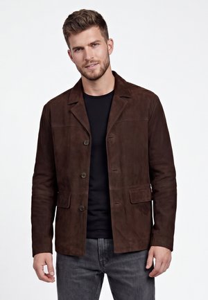Jonge man met kort haar en baard, die een donkerbruine jasje met knopen draagt over een zwart overhemd en grijze spijkerbroek tegen een effen achtergrond.