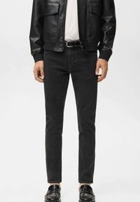 Mann trägt schwarze Slim-Fit-Jeans, schwarze Lederjacke, weißes Hemd, schwarzen Gürtel und schwarze Loafers vor einem schlichten hellen Hintergrund.