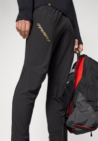 Schwarze, leichte Hose mit einem glänzenden goldenen "Dynafit"-Logo, im taillierten Schnitt und aus strukturiertem Stoff; neben einem schwarz-roten Rucksack gehalten.