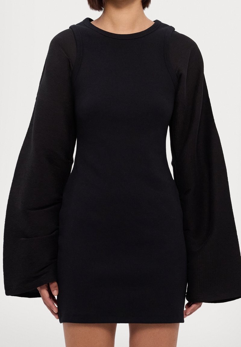 Femme portant une robe noire cintrée avec de longues manches ballon oversize et un col rond sur un fond uni.