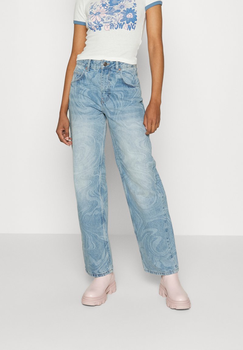Jeans azul claro com um padrão marmoreado, corte solto e cintura alta. Combinados com uma t-shirt branca com detalhes azuis e sapatilhas de plataforma rosa claro.