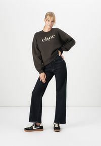 Mujer con sudadera marrón oscura con el texto "choc", jeans anchos oscuros y zapatillas de black y blanco, de pie contra un fondo blanco.