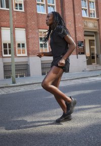 Maglie sportive nere, pantaloni corti leggeri e scarpe da ginnastica texturizzate; postura di corsa su una strada urbana con uno sfondo di mattoni rossi.