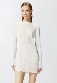 Abito bianco lavorato a maglia con maniche lunghe, motivo a rete aperta e orlo a scallop. La texture è leggera e semi-trasparente.