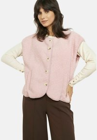 Rozā fleeces veste ar apaļu apakšmali, zelta pogām priekšpusē un sānu kabatām, pārklāta ar krēmkrāsas garām piedurknēm topu.