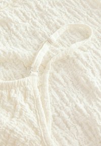 Tissu blanc texturé avec un design froncé. Doté d'une fine sangle avec un curseur en métal argenté pour l'ajustabilité. Doux et léger.