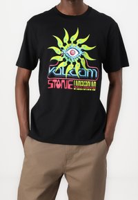 Camiseta de algodón negra con un diseño gráfico colorido que presenta un motivo de sol, un ojo y texto, combinada con pantalones beige. Mangas cortas y cuello redondo.