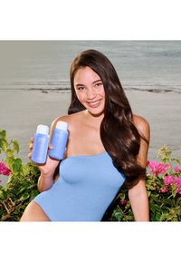 Twee blauwe shampoo-flessen met witte doppen, vastgehouden voor een achtergrond van bloemrijke greenery en een strand. De flessen hebben eenvoudige, moderne etiketten.