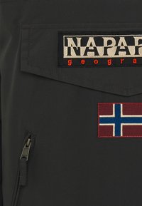 Tela negra con una superficie texturizada que presenta un parche rectangular con "NAPAPIJRI" en beige y rojo, junto a un parche de la bandera noruega en rojo y azul.