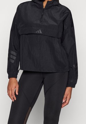 Kvinde iført en sort Adidas vindjakke med delvis lynlås og logo, kombineret med sorte tætsiddende leggings med meshpaneler.