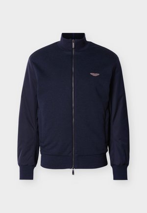 Ναυτικό bomber μπουφάν με ψηλό γιακά, υφασμάτινο σώμα και αντίθετες λείες μανίκια. Διαθέτει φερμουάρ κλεισίματος και μικρή λεπτομέρεια με λογότυπο με φτερά.