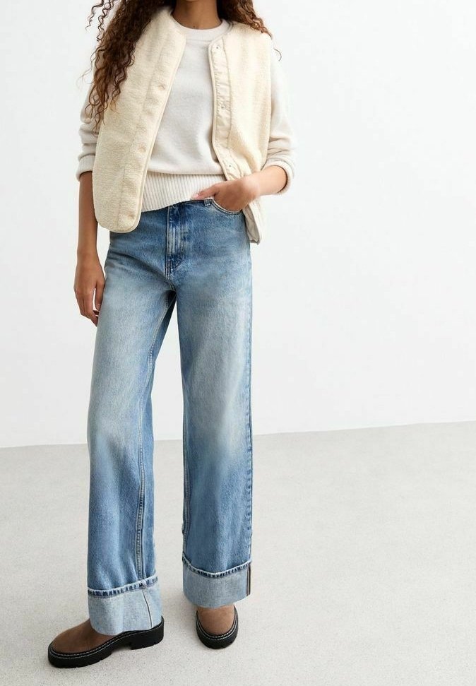 Woman wearing light beige fleece vest over cream sweater, wide-leg cuffed blue jeans, and brown low-heel shoes standing on light floor.