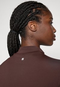 Top atletico marrone con colletto alto, tessuto liscio e un piccolo logo bianco sul retro. Capelli acconciati in trecce, legate alla nuca.