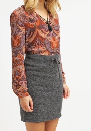 Žena v oranžovém vzorovaném halením s dlouhými rukávy a šedé pletené sukni s vysokým pasem a šňůrkou v pase.