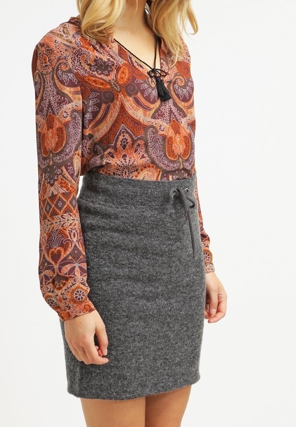 Femme portant un chemisier à manches longues à motif cachemire dans des tons orange et une jupe en maille grise taille haute avec un cordon de serrage à la taille.