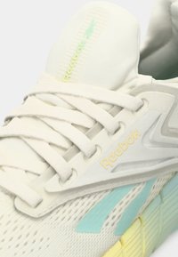 Reebok sportcipők fehér hálós felsőrésszel, textúrált borítással, mentazöld és sárga részletekkel, valamint lapos cipőfűzőkkel a biztos illeszkedés érdekében.