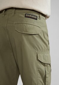 Los pantalones cargo de tela verde oliva cuentan con múltiples bolsillos, costuras reforzadas y un parche con el logotipo en la parte trasera de la cintura.