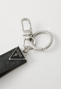 Porte-clés en métal avec une étiquette triangulaire texturée noire ornée d'un logo "GUESS" en argent et d'un point d'interrogation, fixée à un anneau brisé et un clip.