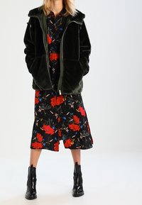 Zelená sametová bunda na zip přes černé květinové šaty s červenými a modrými vzory. Černé kotníkové boty dokončují outfit.