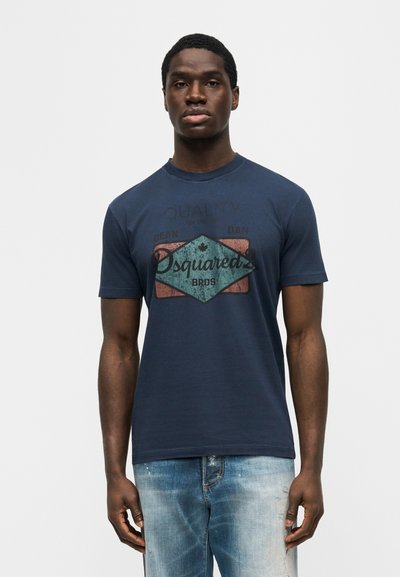 Άνδρας που φοράει μπλε σκούρο t-shirt με λογότυπο Dsquared2 Bros και ξεθωριασμένο μπλε τζιν, στέκεται μπροστά σε απλό ανοιχτό γκρι φόντο.