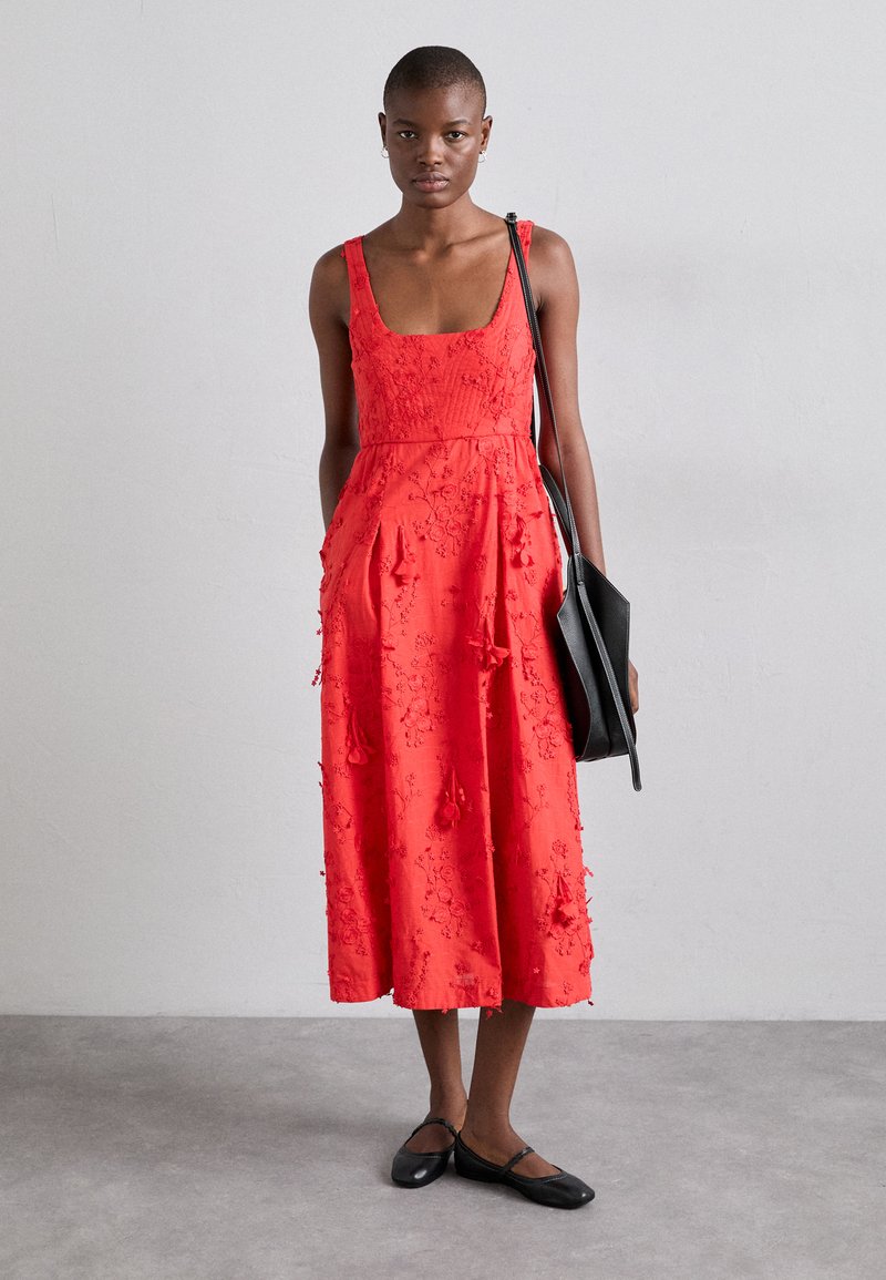 Abito rosso senza maniche con ricami floreali, di lunghezza al ginocchio, dettagli testurizzati e tasche laterali, abbinato a ballerine nere e a una borsa a spalla.