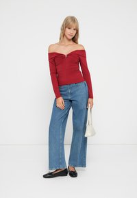 Top rojo de un solo hombro con mangas largas y textura fruncida, combinado con jeans de mezclilla azul de pierna ancha. La modelo sostiene un bolso blanco.