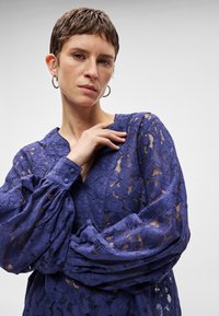 Μωβ δαντελένιο πουκάμισο με V λαιμό, κουμπωτές μανσέτες και φουσκωτά μακριά μανίκια. Διαφανές λουλουδάτο σχέδιο σε όλο το ύφασμα.