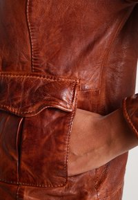 Brun läderjacka med en texturerad yta, framträdande sömmar och en sidoficka. Lädret är mjukt och något slitet.