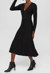Femme portant une robe noire à manches longues avec de fines rayures blanches verticales et des bottines noires pointues à talons.