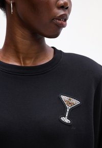 Sudadera negra con un diseño de copa de cóctel decorada con cuentas en el lado izquierdo del pecho, con cuentas transparentes y marrones sobre un fondo de tela texturizada.