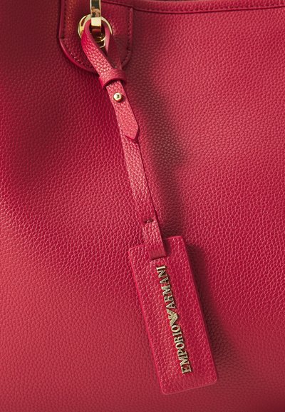 Raudona tekstūruota odinė rankinė su auksiniu furnitūra ir nuimamu žyme, ant kurios užrašyta "Emporio Armani" logotipas. Prisegtas lygus diržas.