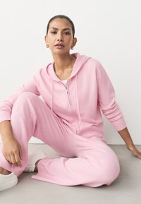 Różowa bluza z kapturem z zamkiem z przodu i ściągaczami na mankietach, w zestawie z dopasowanymi dresami. Gładka faktura, luźny krój i białe trampki.