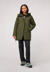 Olijfgroene waterdichte parka met een capuchon, voorzien van een ritssluiting aan de voorkant, zijzakken en een relaxte pasvorm, gedragen met zwarte broek en groene schoenen.
