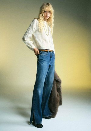 Blusa de manga larga texturizada color beige claro, vaqueros de cintura alta azul con perneras acampanadas y aberturas laterales, cinturón marrón y botas negras de tacón.