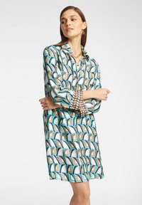 Vestito camicia con un motivo geometrico in turchese, marrone e crema. Dotato di colletto e chiusura a bottoni, con maniche lunghe e vestibilità comoda.