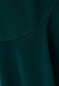 Mörkgrön tyg med en slät textur som har en böjd sömdetalj och subtila veck som skapar mjuka skuggor.