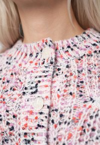 Pletený cardigan z vícetrojného vlákna se žlutými, pomerančovými a fialovými skvrnami. Má kulatý výstřih a velké ozdobné knoflíky.