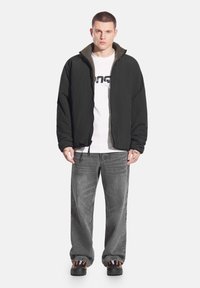 Czarna pikowana kurtka na zamek z miękką zieloną podszewką z polaru, noszona na białej koszulce z nadrukiem, w połączeniu z szarymi szerokimi jeansami i czarnymi butami.