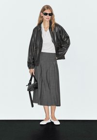 Giacca oversized in pelle nera, blusa bianca, gonna midi plissettata grigia, ballerine bianche, borsa nera, modella che indossa occhiali da sole, sfondo semplice.