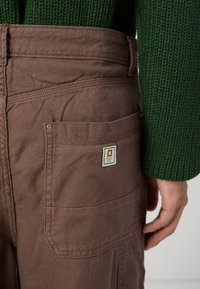 Brown fabric cargo pants with a back pocket and a square logo patch. The texture is sturdy, and the waistband is slightly relaxed.
