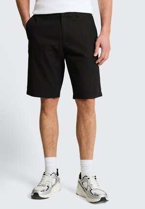 Mann trägt schwarze knielange Shorts, weiße Socken und weiße Sneakers, steht mit einer Hand in der Tasche vor hellem Hintergrund.