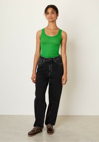 Žalia raštuota liemenė, juodos aukštos juosmens plačios kelnės su priekiniamis kišenėmis ir rudos su raišteliais batai. Neutrali fono spalva ir grindys.