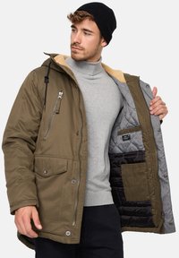 Jachetă izolată de culoare verde măslin cu glugă căptușită cu sherpa, buzunar pe piept cu fermoar și căptușeală matlasată. Include nasturi cu cap și șnururi reglabile.
