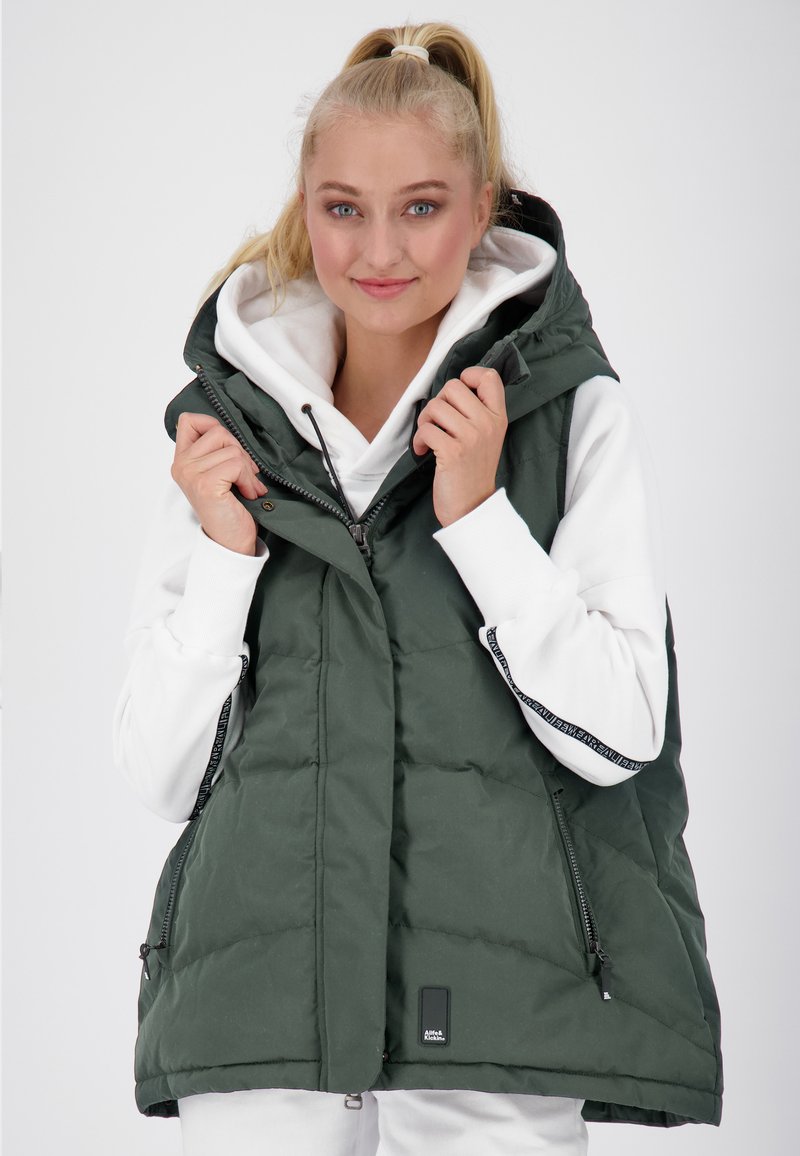 Gilet verde gonfiabile con cappuccio, chiusura zip e tasche laterali; indossato sopra un felpa bianca. Presenta una texture trapuntata e un patch con logo.