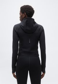 Άτομο που στέκετα ι με την πλάτη στραμμένη, φορώντας μαύρο μπουφάν με κουκούλα και μαύρα κολάν, μπροστά σε απλό ανοιχτόχρωμο φόντο.