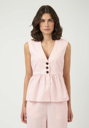 Femme aux cheveux châtains mi-longs portant un haut sans manches rose pâle avec trois boutons noirs et un pantalon assorti sur fond uni.