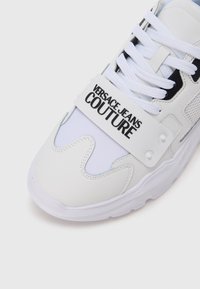 Biały sneaker z gładką skórzaną cholewką, akcentami z siateczki, grubą podeszwą oraz paskiem z napisem "VERSACE JEANS COUTURE".