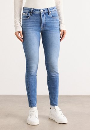 Kobieta ubrana w jasnobeżowe obcisłe dżinsy z delikatnym wyblaknięciem, białą bluzkę z długim rękawem i białe trampki, stojąca na prostym podłożu.